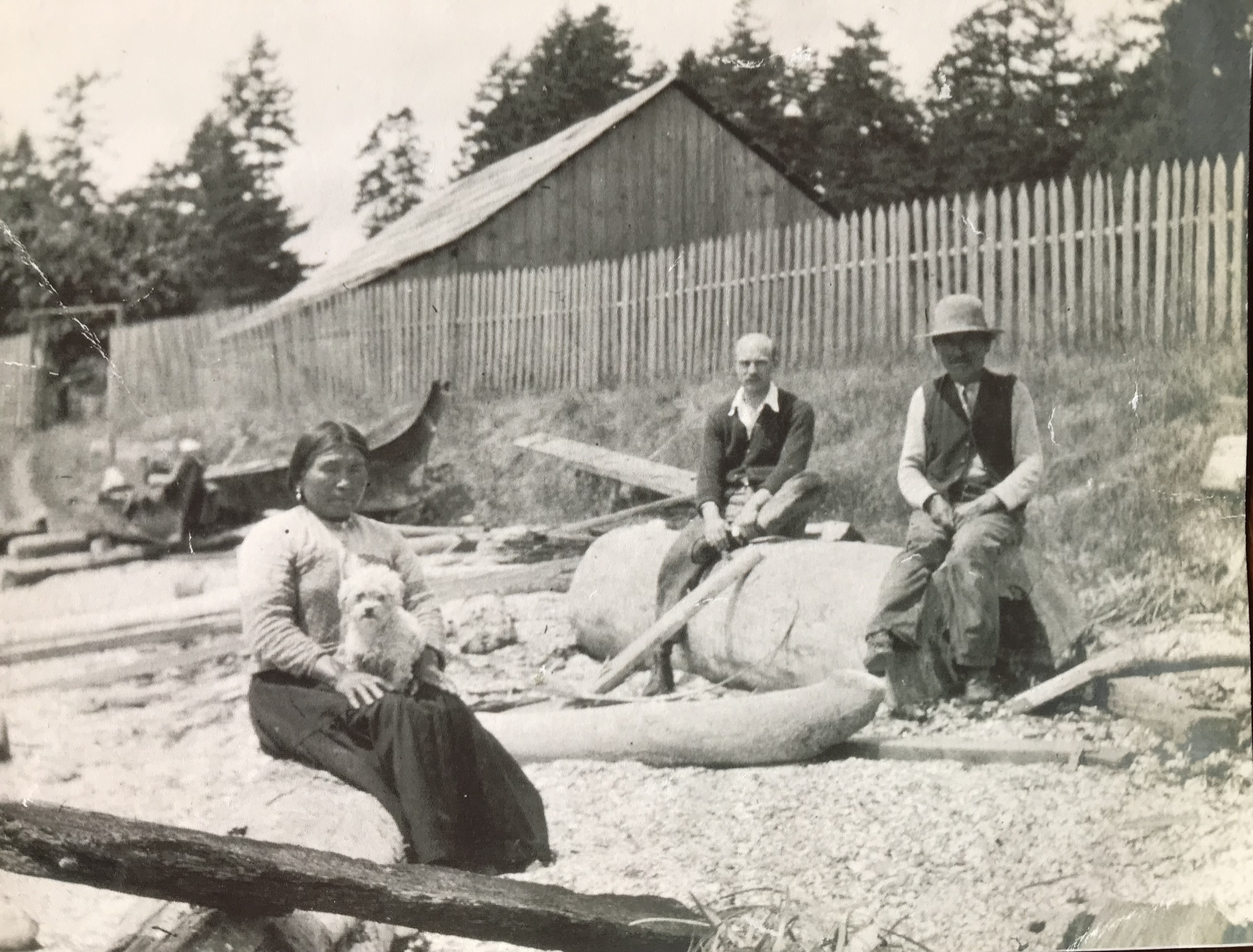 Chief Charlie Sulsulth, his wife Mary and unidentified visitor on the beach in front of the longhouse. Note little dog and Chinook-style canoes.